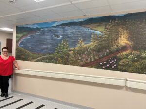 A person stands with a visual art therapy mural on the wall in the background depicting a landscape image with water, flowers and trees.